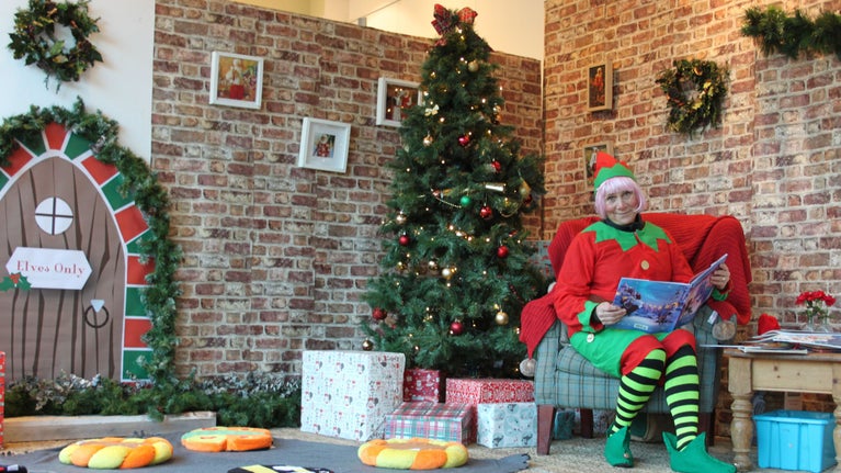A photograph of an person dressed up as an elf sitting on a chair holding a book. The room is decorated in a Christmas theme with a Christmas tree in the centre of the image.